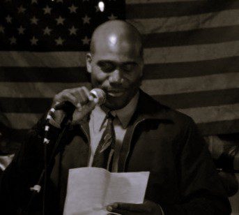 Man reading from a paper into a microphone with an American flag backdrop.